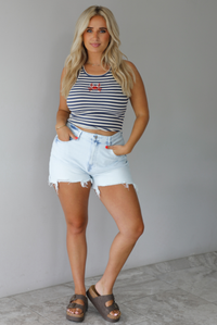 Woman wearing a navy and white striped cropped tank top with beaded crab graphic on the front with light wash denim shorts indoors against a white wall with white marble flooring. 