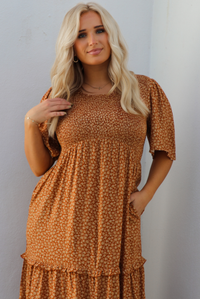 Up close of a woman model outdoors wearing a rust dress with a cream floral print pattern throughout. It has flowy short sleeves, a smocked bodice, and a flowy maxi length skirt with functional pockets on either side. 