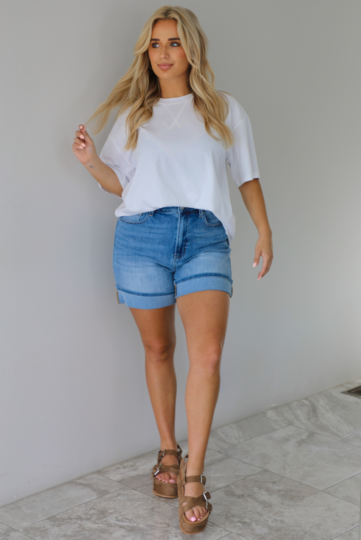 Woman wearing a white oversized tee tucked into medium wash denim shorts indoors against a white wall with white marble flooring. 