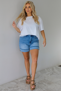 Woman wearing a white oversized tee tucked into medium wash denim shorts indoors against a white wall with white marble flooring. 