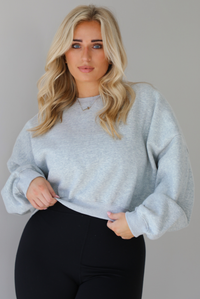 Woman wearing a heather grey cropped sweatshirt with a black fitted jumpsuit underneath. The photo shows a closeup of the sweater where you can really see the heather grey, the cinched details at the wrist and waist, and the cropped cut. The photo is taken indoors against a white wall with white marble flooring. 