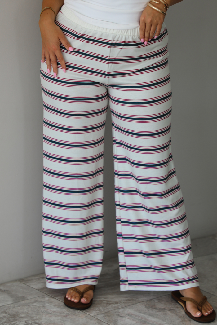A person wearing wide-leg pants with horizontal green and red on a cream-white base, paired with a white top and brown sandals, standing indoors against a white wall with white marble flooring. 