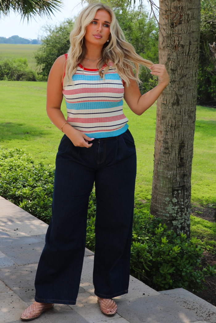 A woman standing outdoors on stone steps next to a tree, wearing a sleeveless striped tank top with red, blue, navy, and white horizontal stripes, paired with dark wide-leg pants and flat sandals.