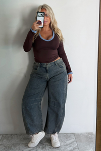 A woman wearing a dark brown, fitted, long sleeved, slightly cropped top with blue trim detailing at the v-neckline and at the sleeve hems, paired with dark acid washed barrel denim jeans, standing indoors in front of a white wall with white marble flooring, taking a mirror selfie. 