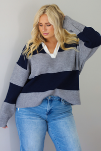 A woman wearing a navy and charcoal boyfriend style pullover top with a white collared detail, with distressed denim, standing indoors against a white wall.
