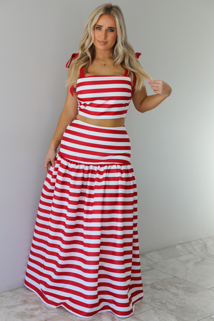 Woman wearing a red and white striped two piece set featuring a cropped top and maxi length, high waisted skirt. The photo is taken indoors against a white wall with white marble flooring. 