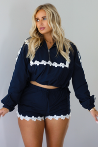A woman modeling a matching navy blue two-piece set with white floral lace trim, consisting of a cropped zip-up jacket and shorts, standing indoors against a white wall.