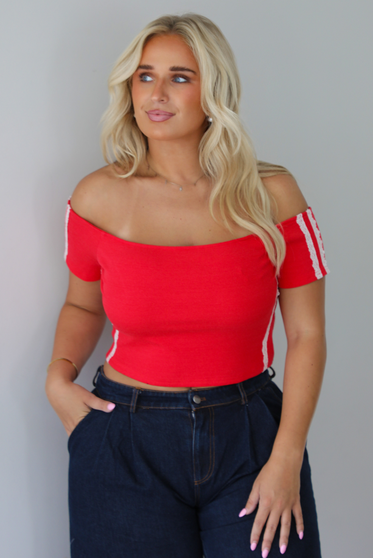 A blonde woman modeling a bright red off-the-shoulder short-sleeve top with white stripes on the sleeves, paired with dark wide-leg denim jeans, standing indoors against a white wall.