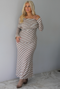 Woman wearing a grey, brown, muted blue, and off white striped fitted maxi dress that has a folded down design at the shoulders and bodice and long sleeves, standing indoors against a white wall with white marble flooring. 