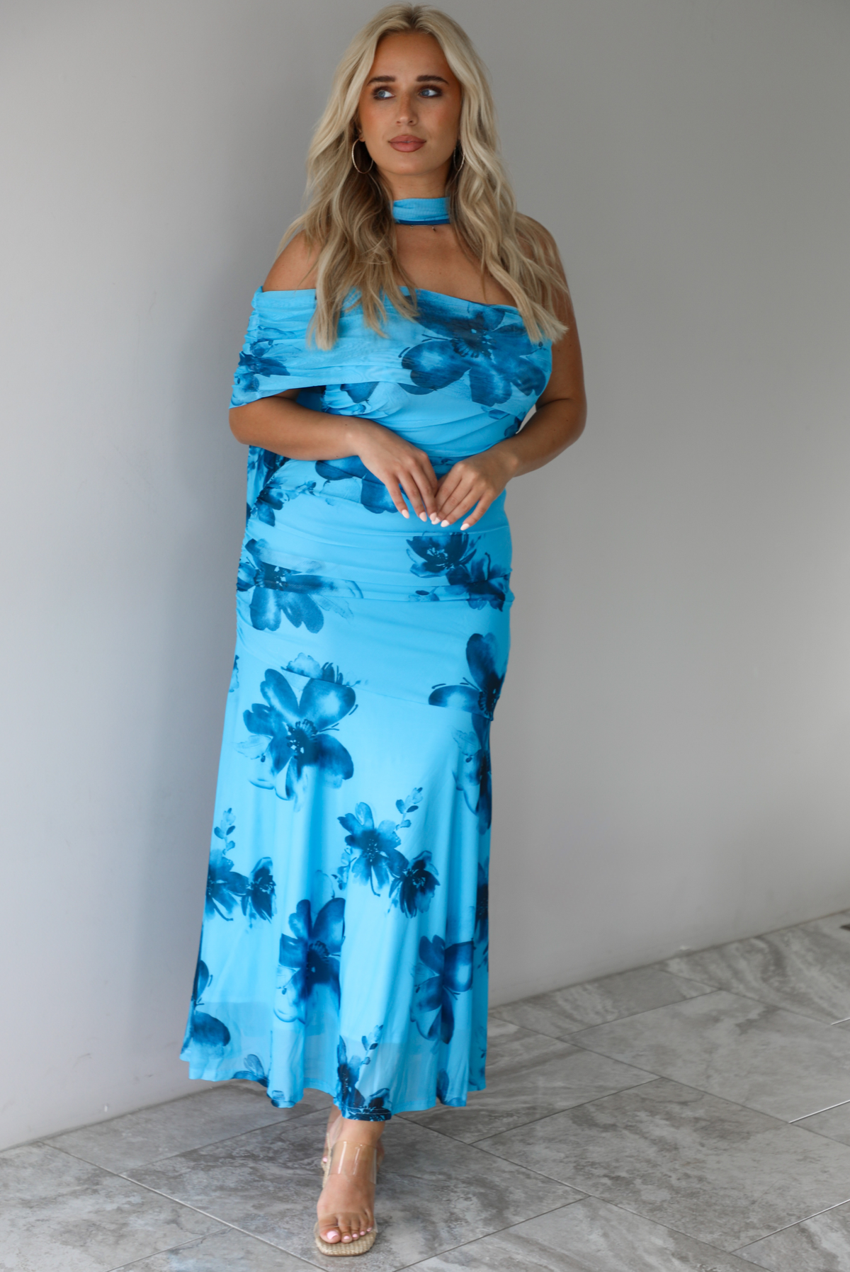 Woman wearing a blue floral maxi dress with a fitted bodice and flowy skirt indoors against a white wall with white marble flooring. 