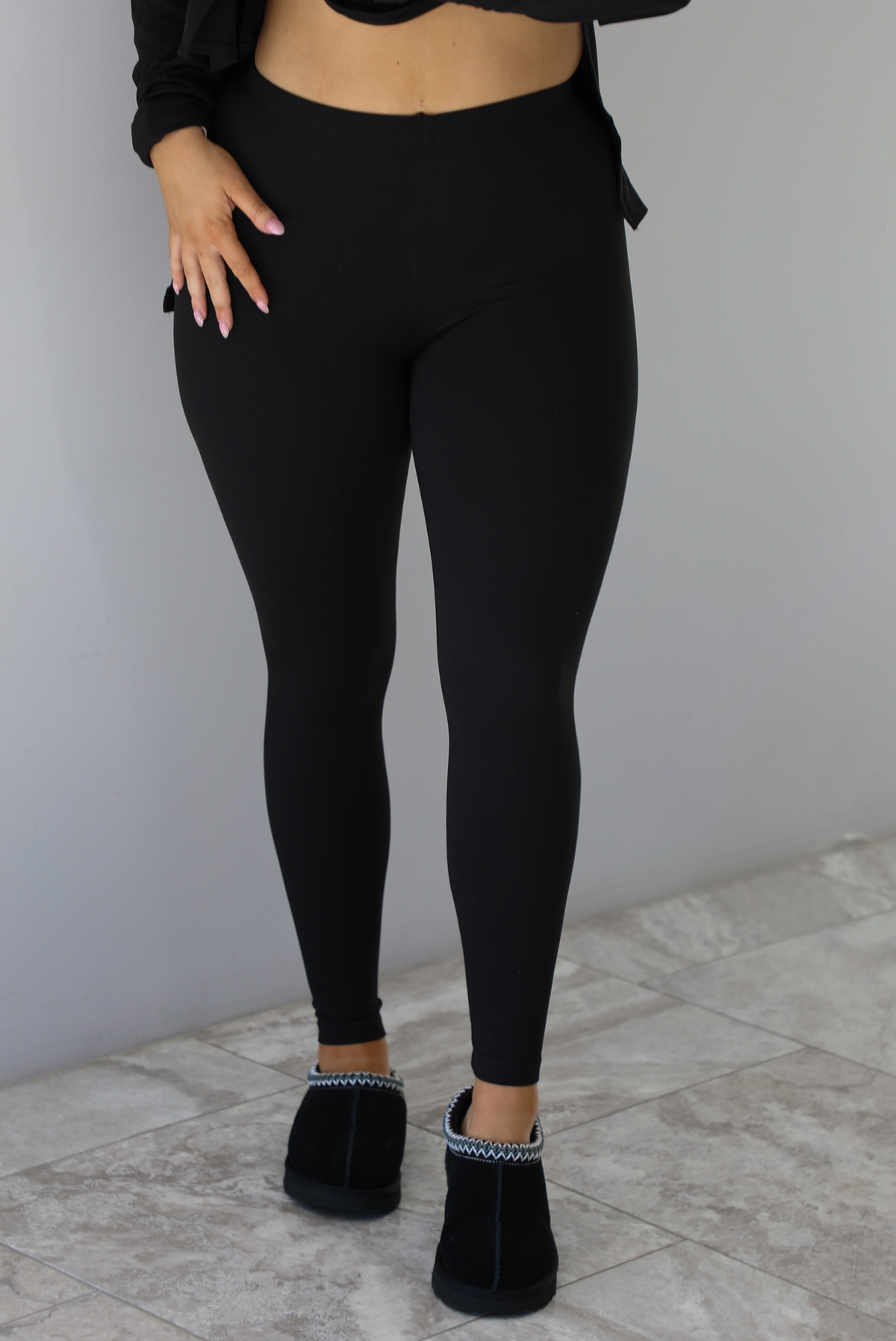 Woman wearing a black two piece set that features an oversized long sleeved top and fitted leggings. The photo shows a closeup of the leggings. The photo is taken indoors against a white wall with white marble flooring. 