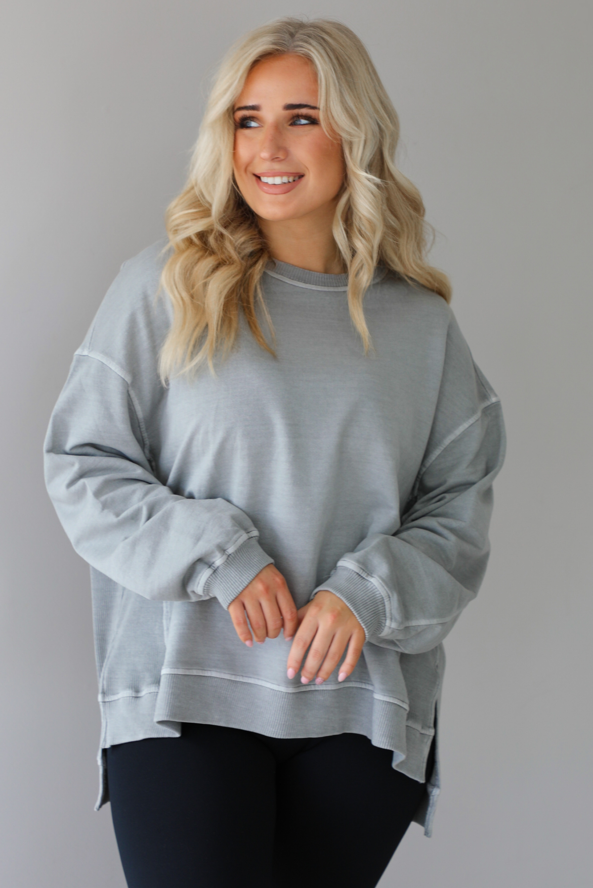 Woman wearing a grey oversized sweater with black leggings that have a slightly flared hem with split hem details in the front. The photo shows a closeup of the sweater and has a white background. 