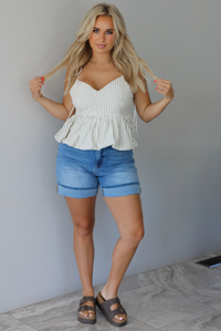 Woman wearing a white and taupe striped tank with medium wash denim shorts indoors against a white wall with white marble flooring. 
