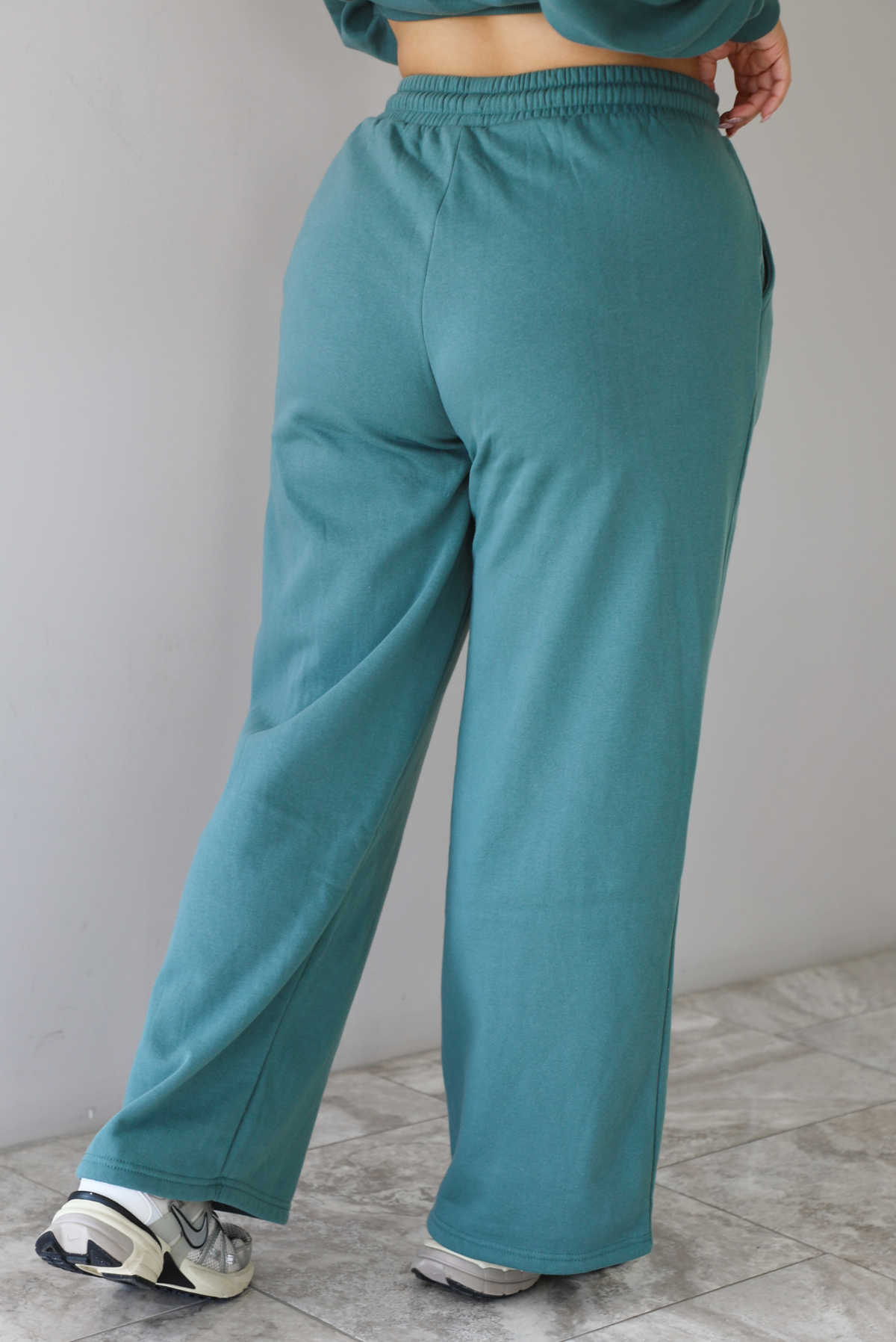Woman wearing a sage two piece sweat-set. The set features a slightly oversized sweater and high waisted sweat pants that have an elastic and drawstring waistline with pockets.The photo shows a closeup of the back of the pants. The photo is taken indoors against a white wall with white marble flooring. 