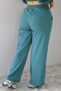Woman wearing a sage two piece sweat-set. The set features a slightly oversized sweater and high waisted sweat pants that have an elastic and drawstring waistline with pockets.The photo shows a closeup of the back of the pants. The photo is taken indoors against a white wall with white marble flooring. 