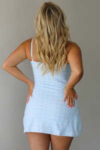 A woman wearing a light blue and white plaid mini dress, standing indoors against a white wall. The photo shows the back of the dress, which has adjustable straps. 