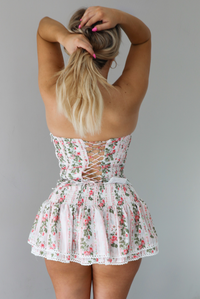 Woman wearing a white and pink floral two piece set featuring a strapless corset style top and an a ruffled skirt. The photo shows a closeup of the back of the set, with the lace up detail in the back of the top. The photo is taken indoors against a white wall.
