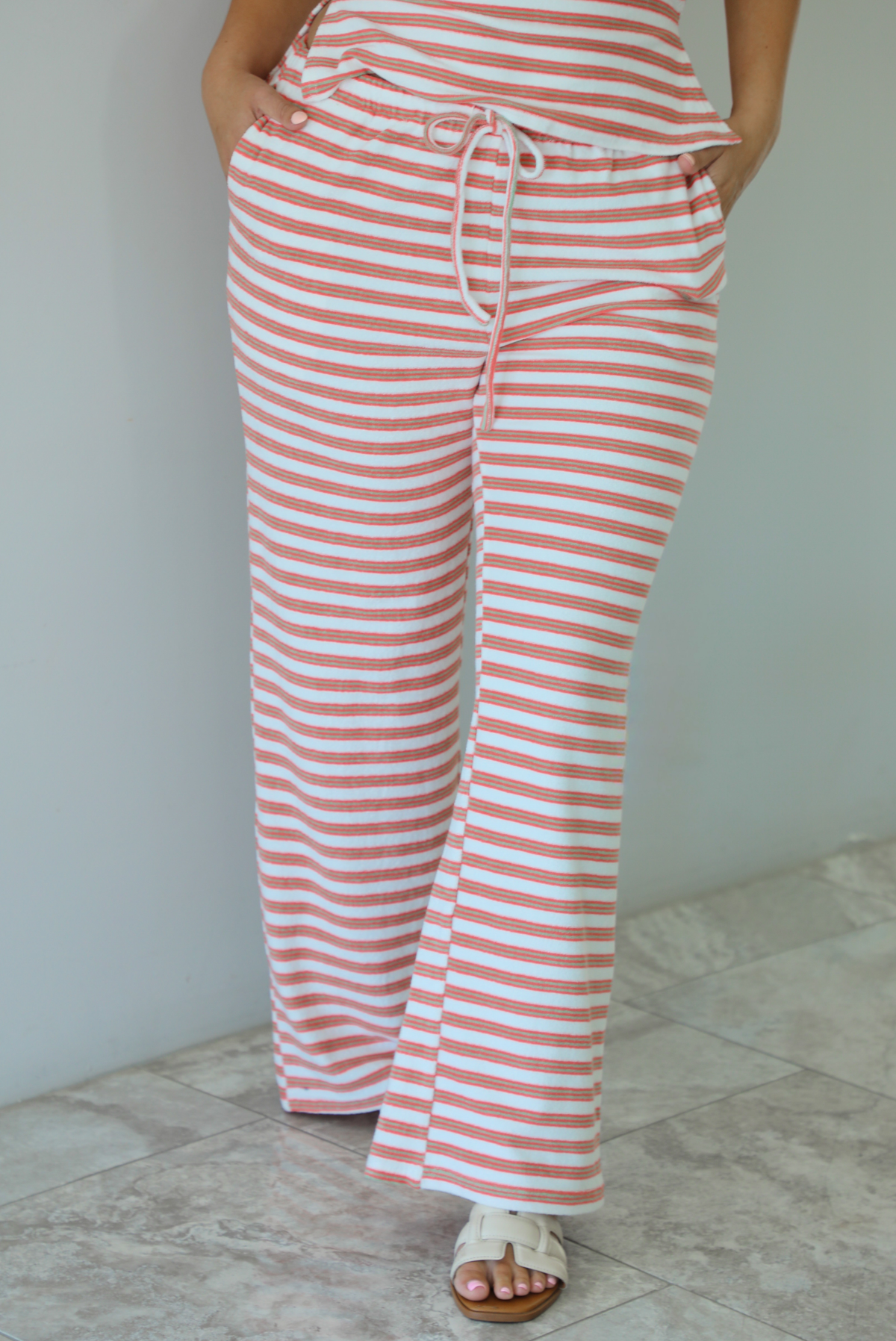 Woman wearing an orange, white, and green striped pattern set that has a strapless tube top and high waisted pants indoors against a white wall with white marble flooring and shows a closeup of the pants, which have an elastic and drawstring waistline with functional pockets on either side. 