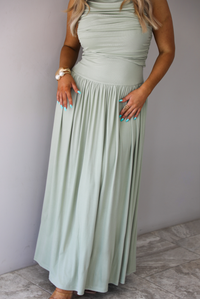 Woman wearing a sage green maxi length dress, which has a rouched detail at the bust, a fitted waistline, a flowy skirt. The photo is taken indoors against a white wall with white marble flooring. 