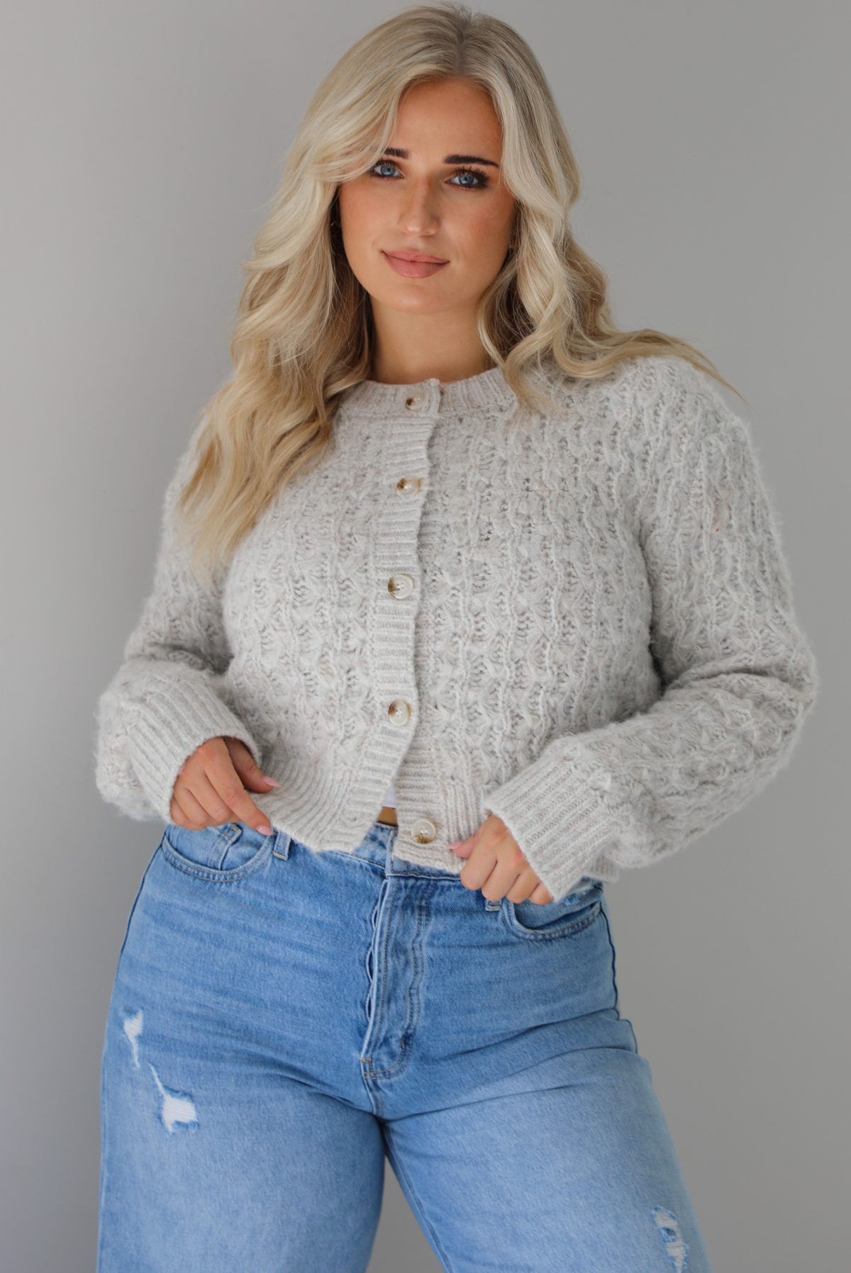 Woman wearing a light grey cardigan buttoned up with high waisted flared hem medium wash denim. The photo shows a closeup of the texture of the cardigan, which resembles a loose textured cable knit. The photo is taken indoors against a white wall.