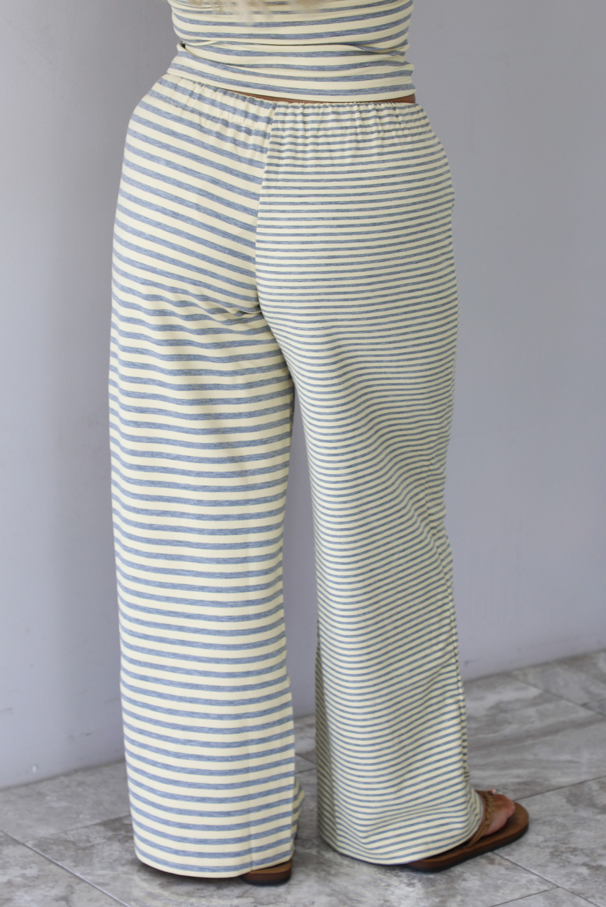 A woman wearing a pale yellow and heather grey striped two piece set with a sleeveless tube top and wide-leg pants, focused on the back of the pants, which have a drawstring waistline, standing indoors against a white wall with white marble flooring. 