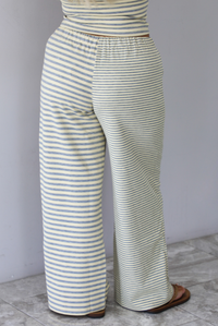A woman wearing a pale yellow and heather grey striped two piece set with a sleeveless tube top and wide-leg pants, focused on the back of the pants, which have a drawstring waistline, standing indoors against a white wall with white marble flooring. 