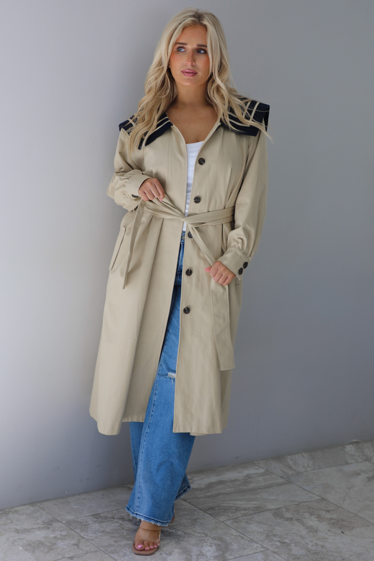 A woman wearing a tan trench coat with a navy collared neckline, functional button closures in the front, functional pockets on either side, with a tie waistline, paired with light wash flared hem denim jeans, standing indoors against a white wall with with white marble flooring. 