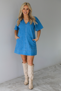 Woman wearing a v-neck, short sleeved, denim mini dress with functional pockets on either side. The photo is taken indoors against a white wall with white marble flooring. 
