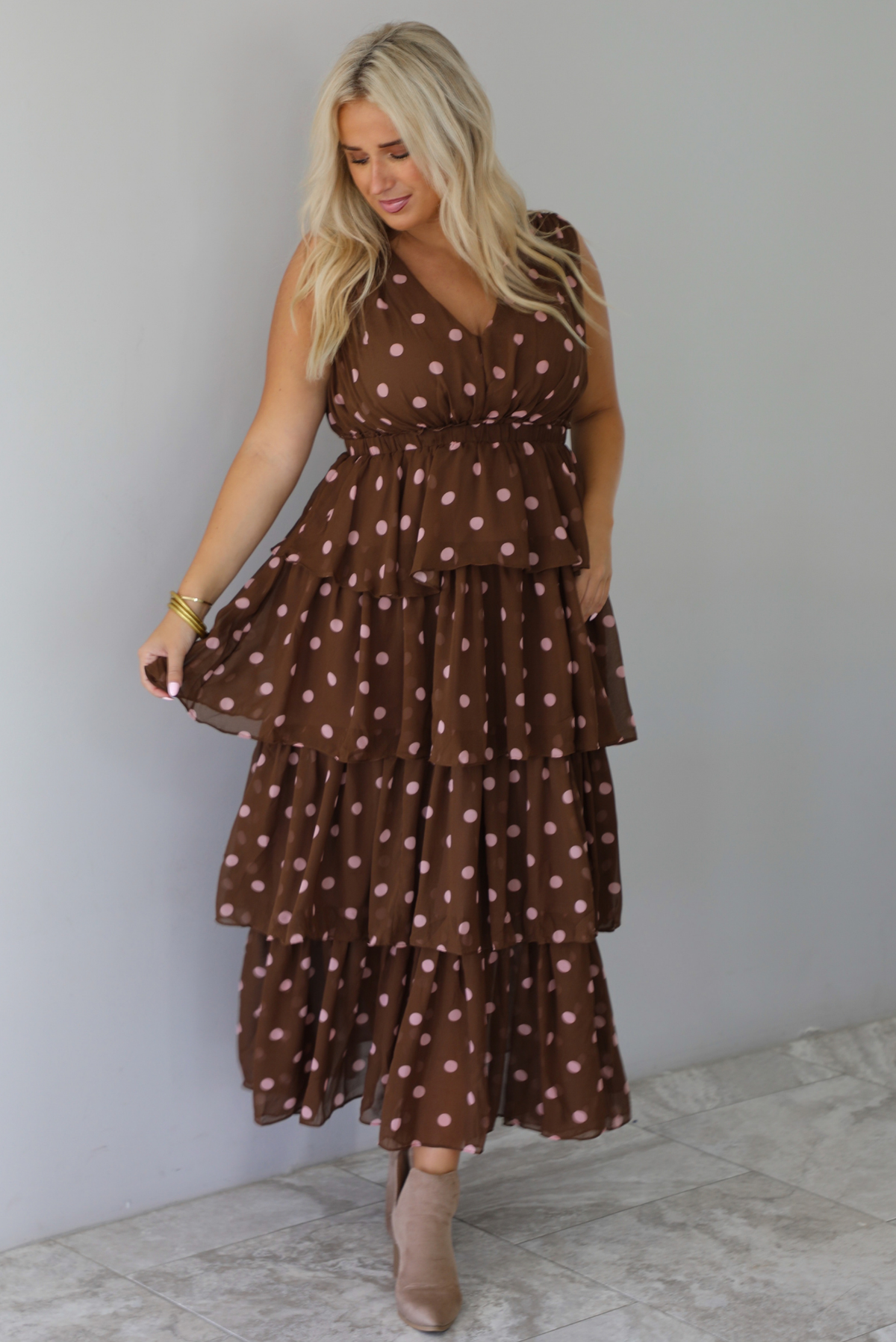 A woman wearing a brown midi length ruffled dress with a pink polka dot print pattern throughout, standing indoors against a white wall with white marble flooring. 