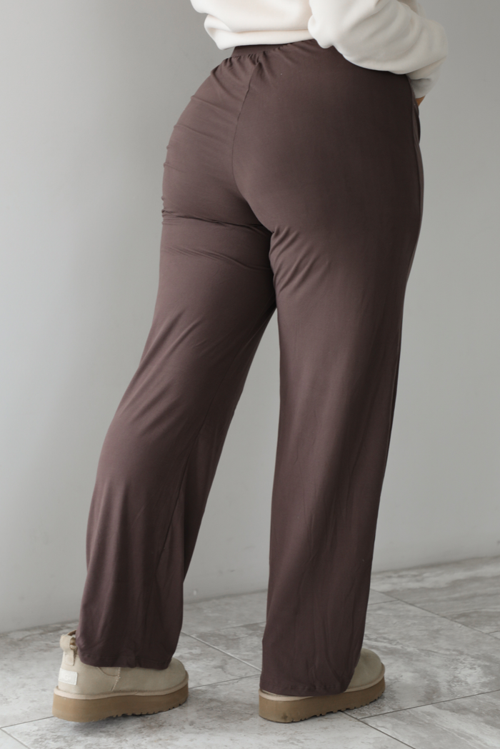 The photo features a model wearing high waisted brown pants that have an elastic and drawstring waistline, a slightly flared hem, and are super soft and stretchy. They are worn with beige ugg boots. The photo is taken indoors against a white wall with white marbled flooring. The photo shows the back of the pants. 