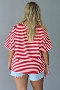 A woman modeling a red and white horizontally striped short-sleeve t-shirt with light blue denim shorts and brown sandals, standing indoors against a white wall. The photo shows the back of the tee. 