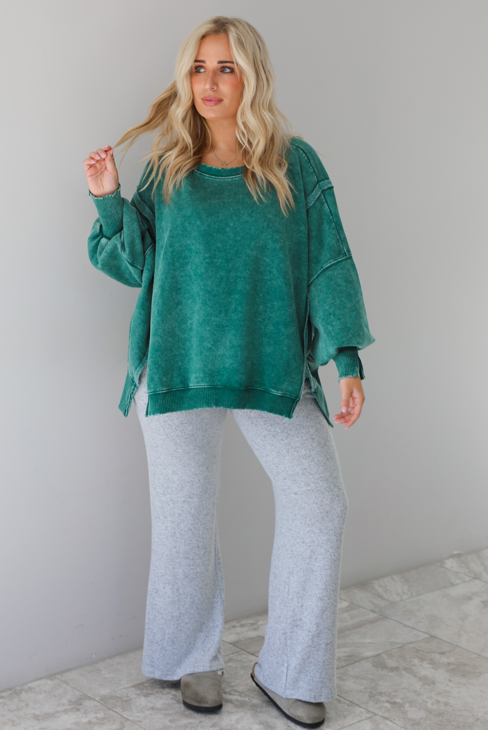Woman wearing green colored oversized sweatshirt with exposed seam details and a high-low design with heather grey casual pants. The outfit is styled with grey slip on shoes. The photo is taken indoors against a white wall with white marble flooring. 