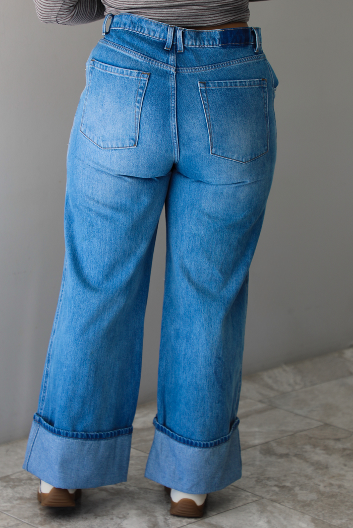 A closeup of a woman shown from behind wearing high waisted medium wash denim jeans with a cuffed hem, standing indoors against a white wall with white marble flooring. 