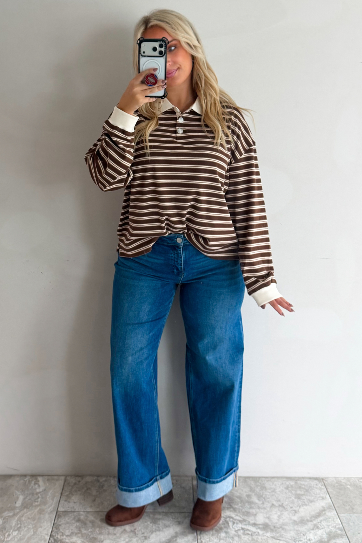 A woman taking a mirror selfie wearing a brown and cream striped collared quarter-button top, paired with dark, wide leg denim with a cuffed hem, standing indoors against a white wall with white marble flooring. 