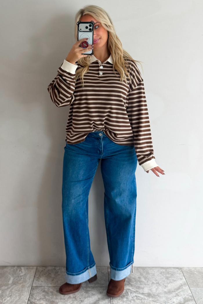 A woman taking a mirror selfie wearing a brown and cream striped collared quarter-button top, paired with dark, wide leg denim with a cuffed hem, standing indoors against a white wall with white marble flooring. 