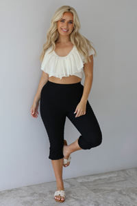 Woman wearing a cream ruffled cropped top with high waisted black, textured capri pants indoors against a white wall with white marble flooring. 