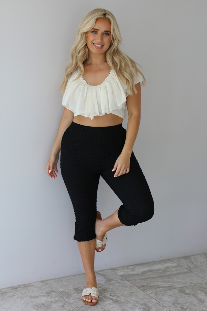 Woman wearing a cream ruffled cropped top with high waisted black, textured capri pants indoors against a white wall with white marble flooring. 