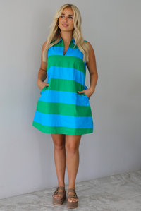 A woman wearing a sleeveless collared mini dress with bold horizontal stripes in green and blue, standing indoors against a white wall with white marble flooring. 
