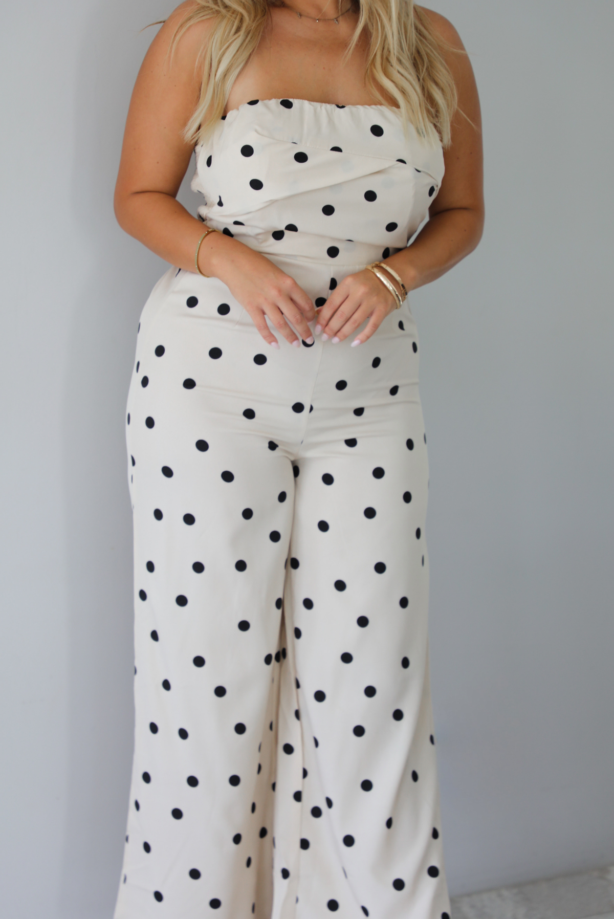 A woman wearing a sand strapless jumpsuit with a black polka dot print pattern throughout, standing indoors against a white wall. The photo is focused on the jumpsuit itself, not showing the models face. 
