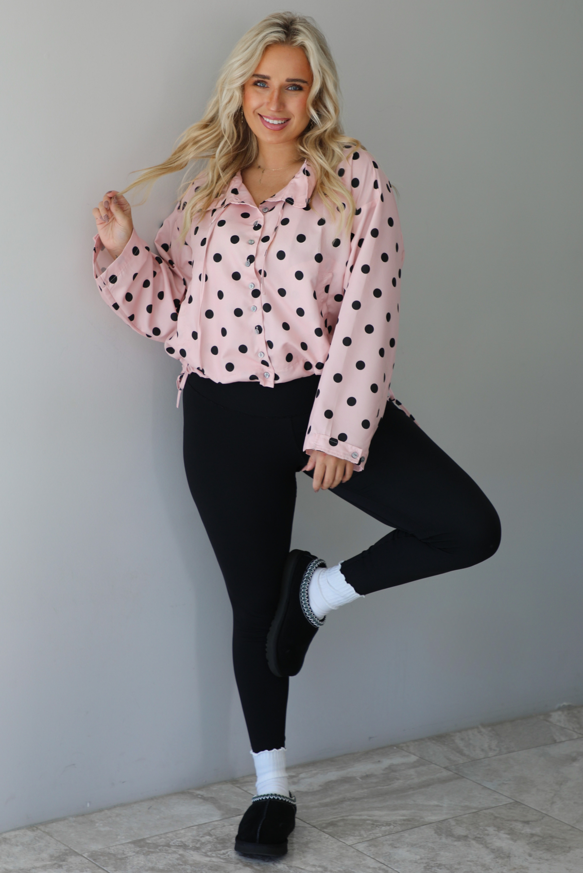 A woman wearing a pink button-down jacket with a black polka dot print pattern, paired with black leggings, white ankle socks, and black slide on slippers, standing indoors against a white wall with white marble flooring. 