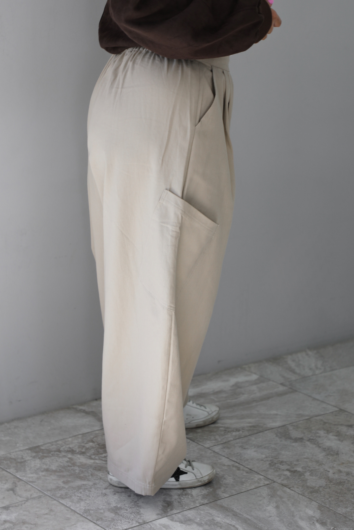 A woman shown from the waist down from the side wearing a pair of sand baggy pants with functional pockets on either side and more pockets further down on the thigh just above the knees, standing indoors against a white wall with white marble flooring. 