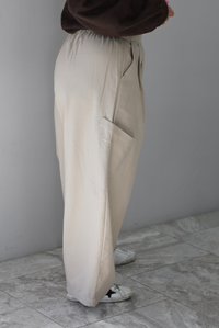 A woman shown from the waist down from the side wearing a pair of sand baggy pants with functional pockets on either side and more pockets further down on the thigh just above the knees, standing indoors against a white wall with white marble flooring. 