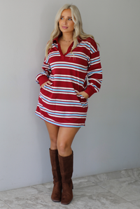 A woman wearing a red burgundy mini dress with a white and blue striped pattern throughout, collared neckline, long sleeves, and functional pockets on ether side, standing indoors against a white wall with white marble flooring. 