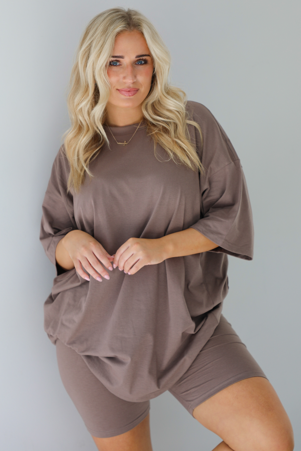 Woman wearing a two piece taupe set featuring an oversized t-shirt and fitted biker shorts.  The photo is taken indoors in front of a white wall.