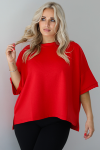 Woman wearing an oversized red t-shirt with black, fitted lettings. The photo shows a closeup of the red top that has split hem details on either side and is slightly longer in the back. The photo is taken indoors against a white wall.