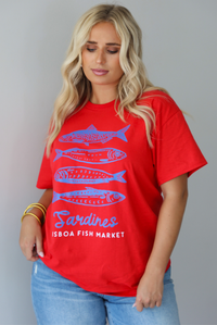 A woman wearing a red graphic t-shirt with blue sardine illustrations and the text 'Sardines LISBON FISH MARKET', paired with blue jeans, standing indoors against a white wall.