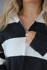 Woman wearing a charcoal and white striped quarter-zip sweater with a collared neckline, raw seam details, and a relaxed fit. It's paired with a fitted black jumpsuit. The photo shows a close up of the quarter-zip. The photo is taken indoors against a white wall with white marble flooring.