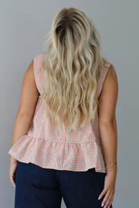 A woman shown from behind wearing a light maroon and cream gingham flowy top with floral details at the straps of the tank, paired with dark denim jeans, standing indoors against a white wall.
