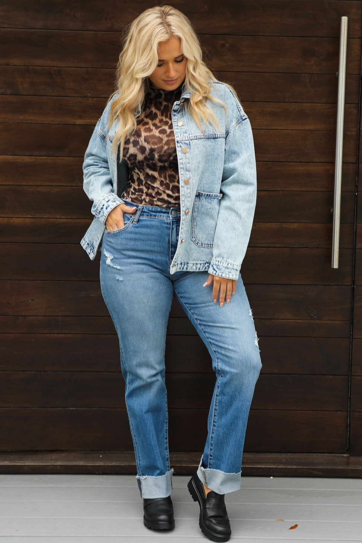 Woman model wearing an oversized, acid washed, light denim jacket, paired with a sheer mesh leopard print top, medium wash denim jeans, and black shoes. The photo is taken outdoors against a dark brown door. The model is standing with a side of the jacket pushed back to reveal more of the top with their hand tucked into the right front pocket of the denim jeans. 