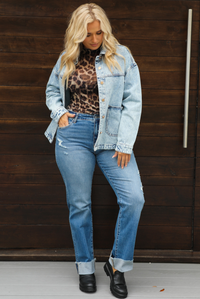 Woman model wearing an oversized, acid washed, light denim jacket, paired with a sheer mesh leopard print top, medium wash denim jeans, and black shoes. The photo is taken outdoors against a dark brown door. The model is standing with a side of the jacket pushed back to reveal more of the top with their hand tucked into the right front pocket of the denim jeans. 